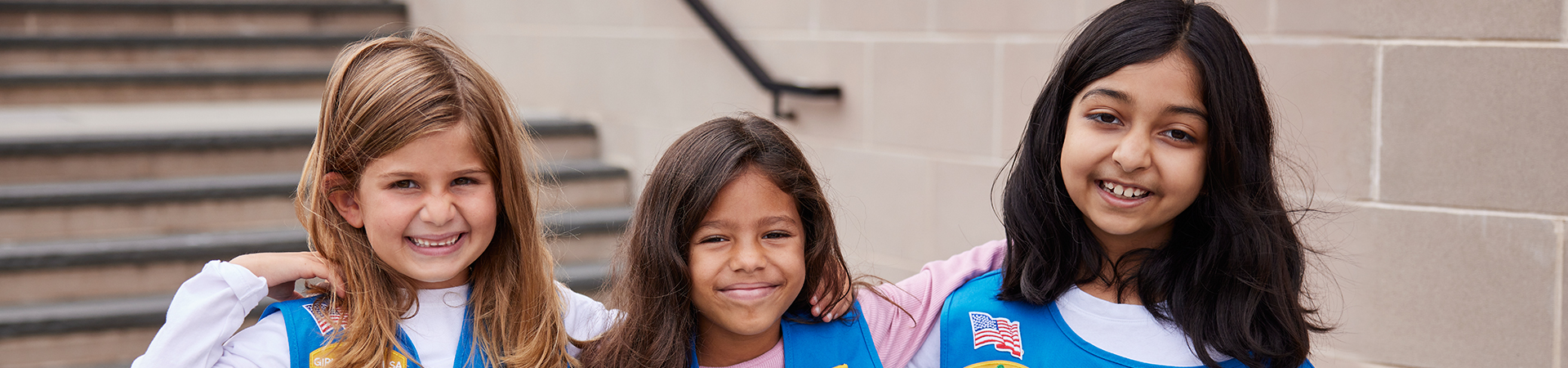  two girl scouts wearing trefoil clothing leaning on one another indoors 